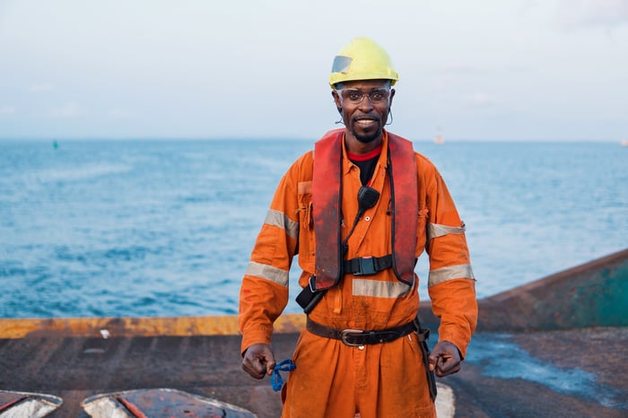 A cheerful man wearing orange high-visibility overalls with reflective stripes and a yellow safety helmet. He is equipped with a black harness, presumably for safety, and stands confidently on a ship's deck with the open sea and a clear blue sky in the background