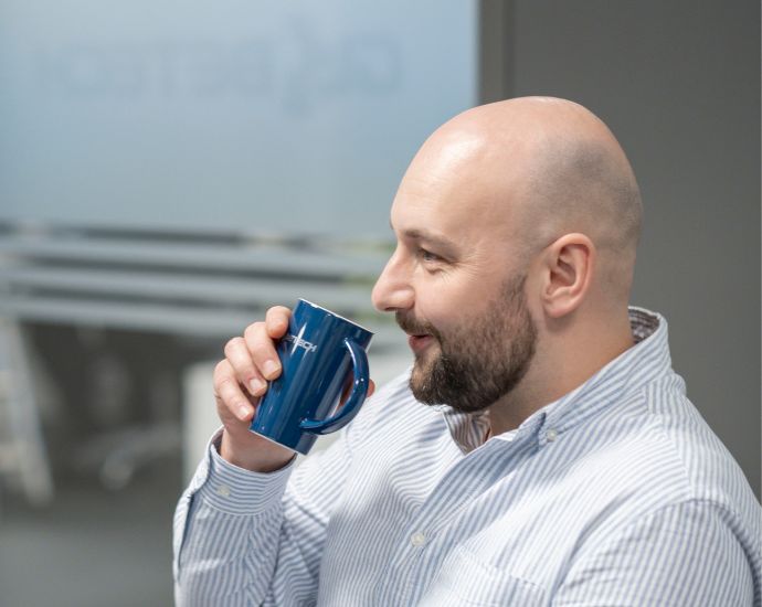 A profile view of a bald man with a beard, wearing a striped shirt, taking a sip from a blue mug in an office setting